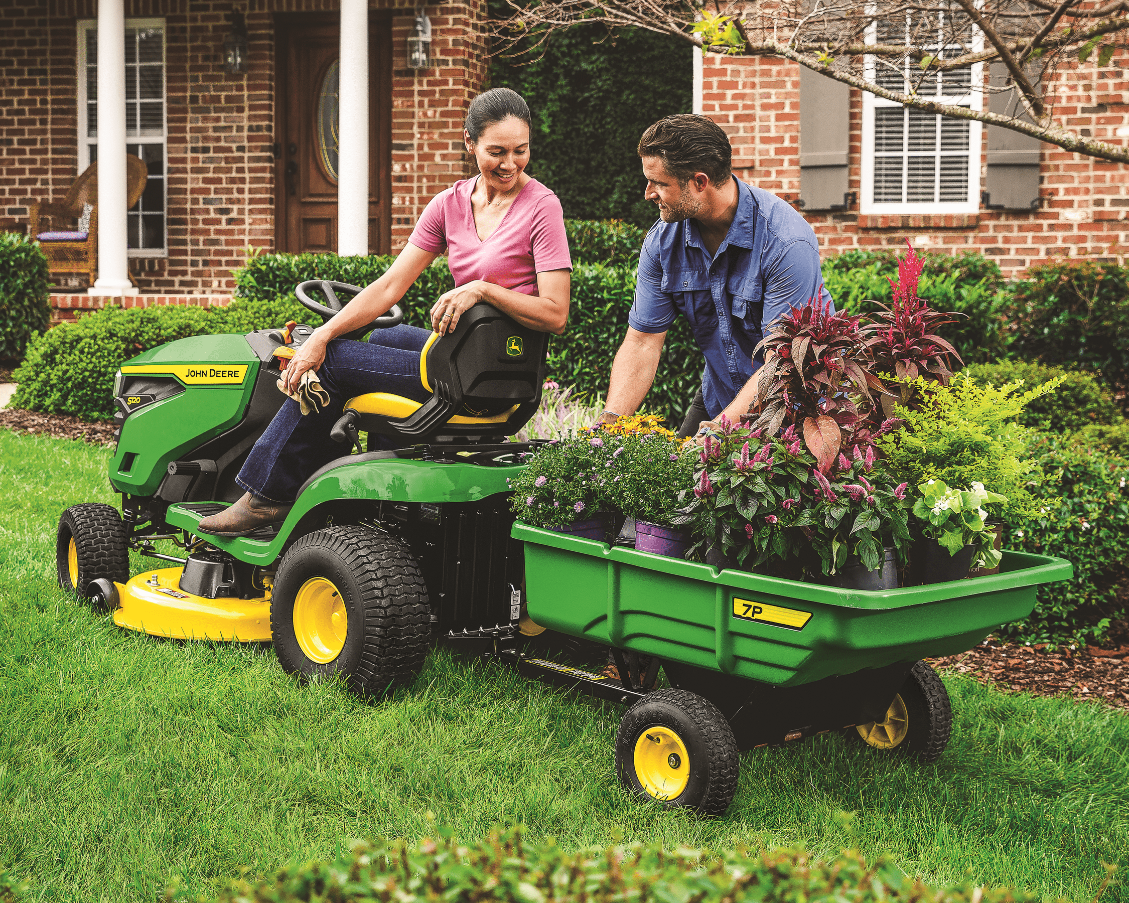 A couple is doing gardening work using a John Deere s120 lawn tractor with 7p cart. 